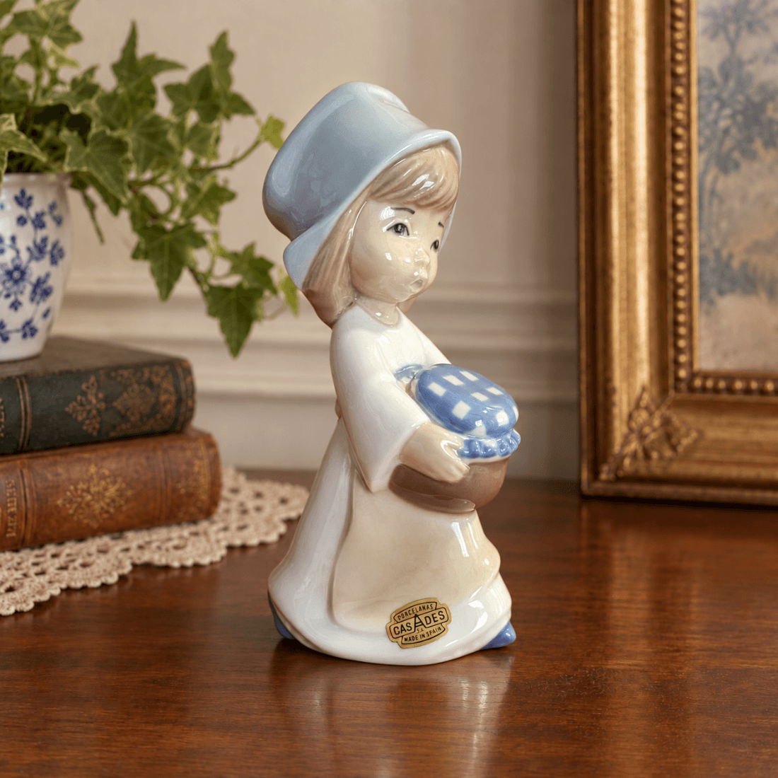 Casades porcelain girl holding a covered jar, displayed on a wooden surface beside books and a framed picture