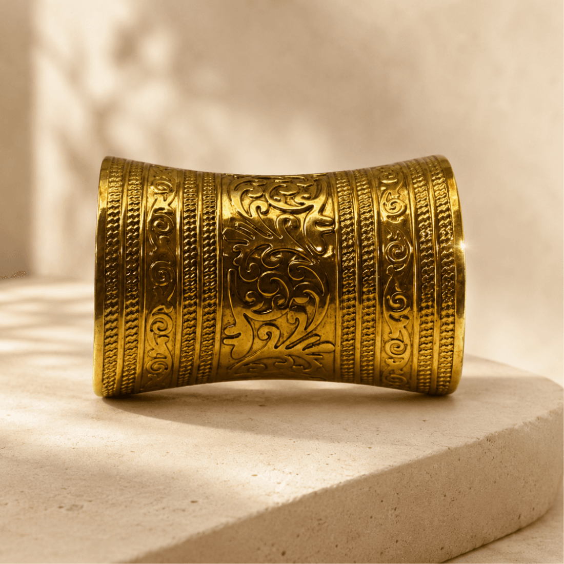 Gold-tone embossed cuff bracelet displayed on a pale round plinth in warm light