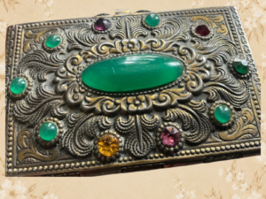 Vintage ornate metal jewellery box with a green central stone and coloured decorative stones on a floral background.